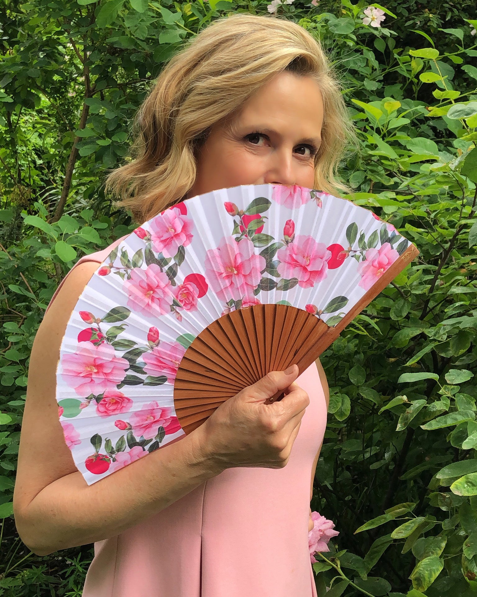 Liz Earle holding Khu Khu X Liz Earle Beautiful Cool Camellias Hand-Fan with collaboration print (Floral with pink and green spots and camellias.)