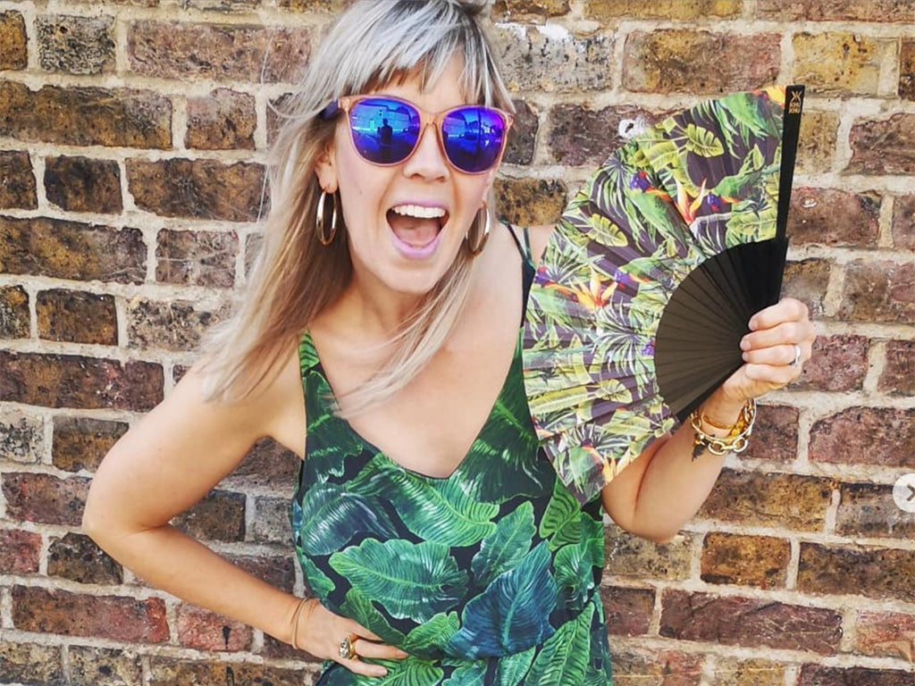 Blogger wears tropical jumpsuit and sunglasses in front of a wall holding Khu Khu tropical Green Parakeet hand-fan. Beautiful green birds perch amongst tropical leaves and orange passion flowers. Fan mounted with black painted wood sticks and mounted with high grade cotton. 