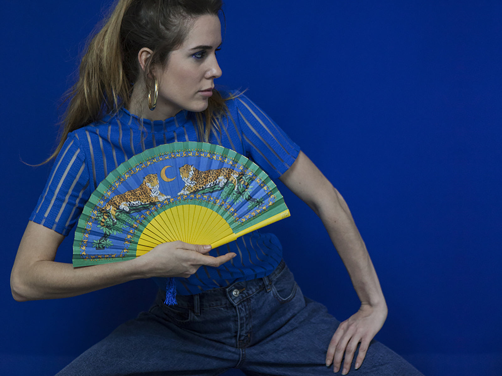 Model wearing blue on background using Khu Khu Lazy Leopards Cool Cats Hand-Fan. Two leopards sit in branches with blue night sky in background mounted with painted yellow sticks with loop and blue tassel.  